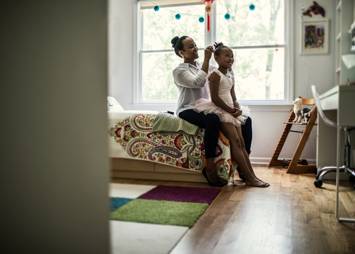 Sitting on edge of a bed, mom arranging hair of daughter who is dressed as a ballerina. 